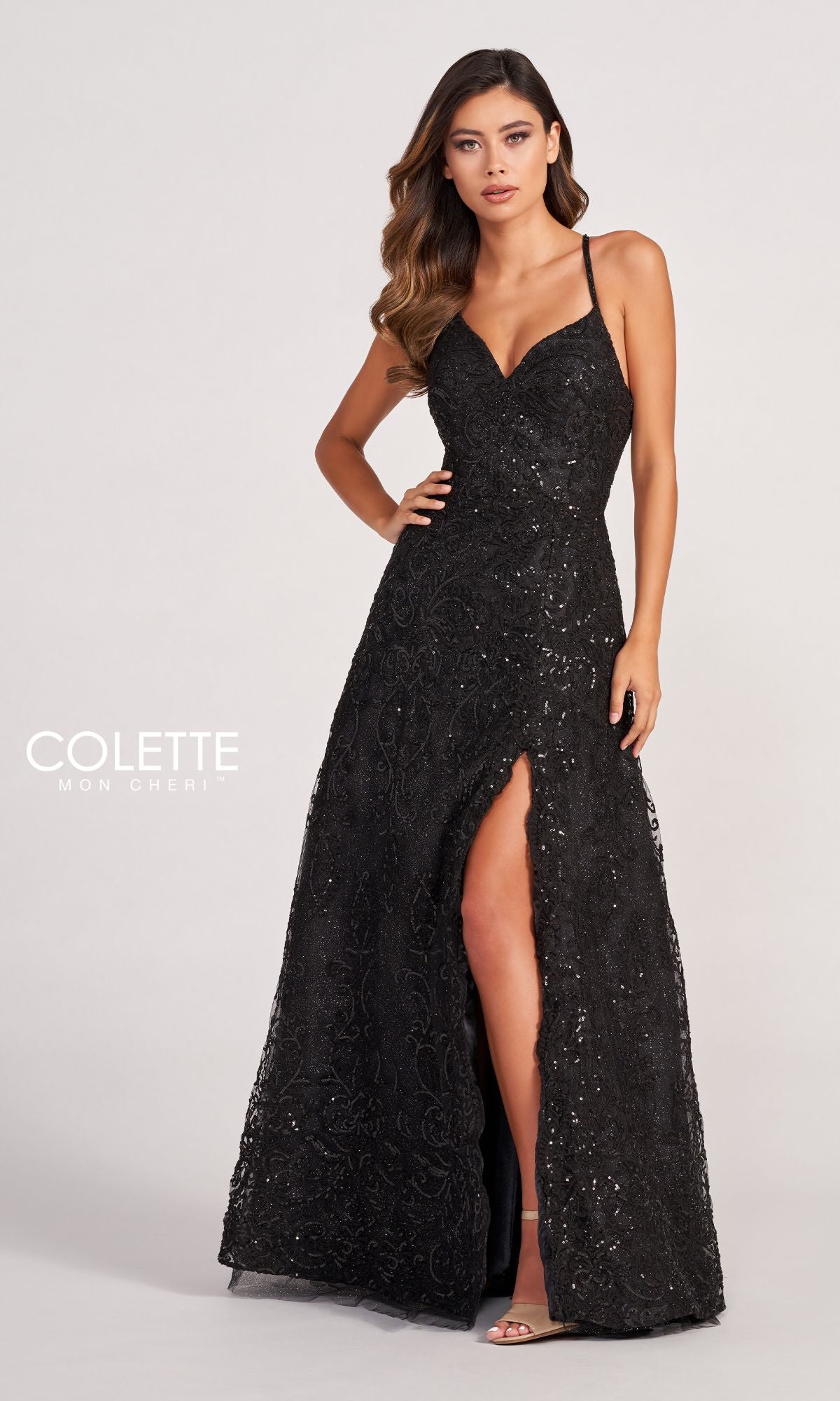 A woman poses against a plain light background in the Colette CL2028 Strappy-Back Lace A-Line Prom Dress—black, floor-length, with thin straps, open back, high slit, fitted bodice, and subtle floral lace patterns.
